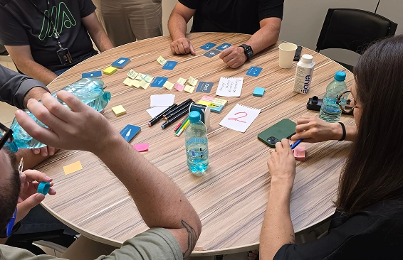 Een groep mensen zit rond een ronde houten tafel tijdens een brainstormsessie met plaknotities, pennen en flessen water